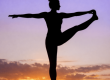 Silhouette of a person balancing in a yoga pose on one leg at sunrise, with the sun low on the horizon and a gradient sky shifting from orange to purple.